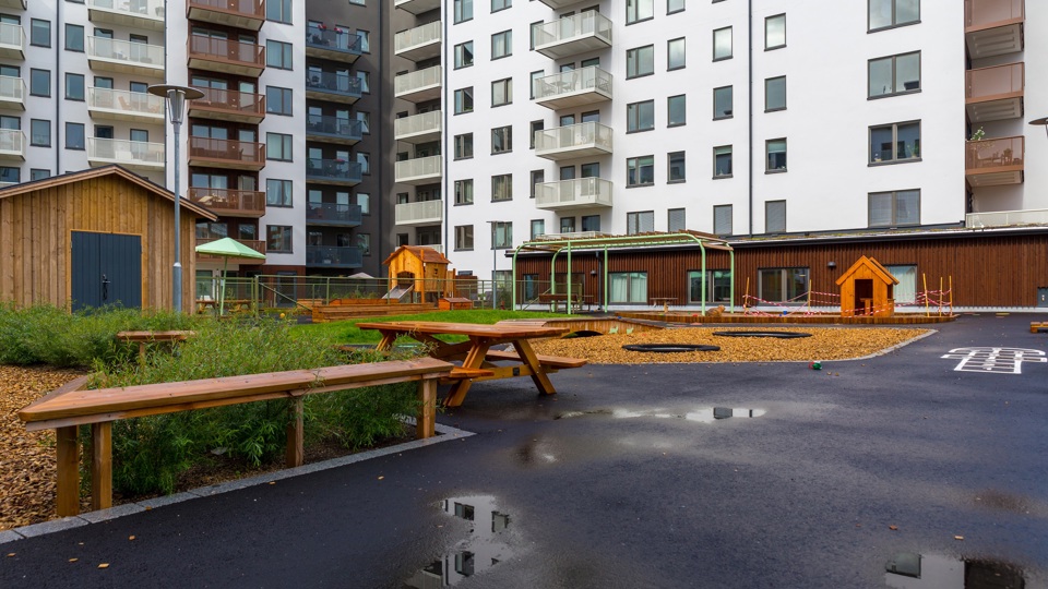 Innergård till flerbostadshus med asfalt, buskar och lekobjekt.