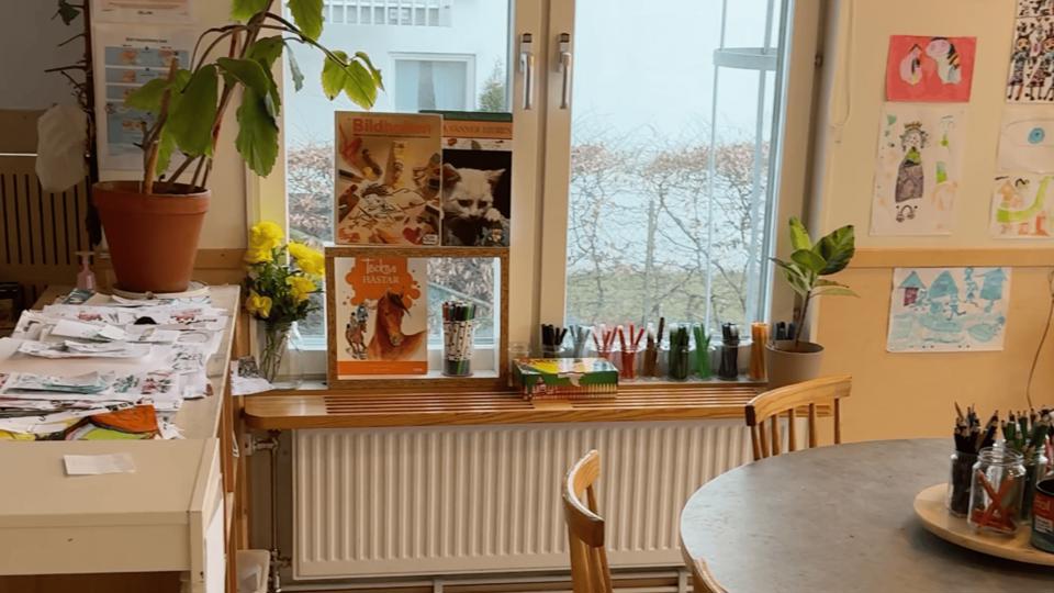 Inomhusvy med fönsterbräda fylld med böcker, pennor och blommor. Bord med papper och konstmaterial.