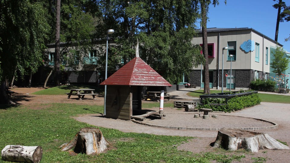 En grön gård med stubbar och lövträd. En lekstuga i trä och rött tak står på gården framför en beige förskolebyggnad i två plan. Byggnaden har brunt tegel i nederkant och mintgröna balkonger, fönsterkarmar och detaljer.