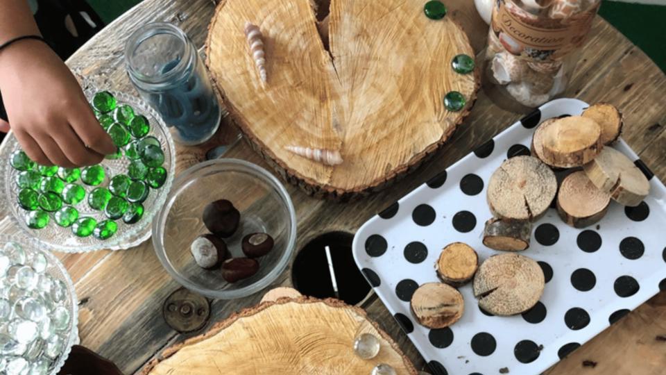 En hand som placerar gröna glasstenar på träbitar och dekorationer som snäckor och kastanjer på ett bord i en förskolemiljö.