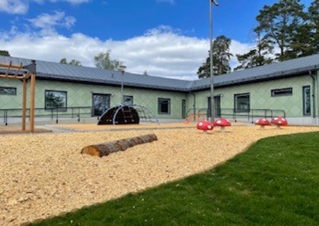 Grön förskolebyggnad bakom en lekyta med sågspån