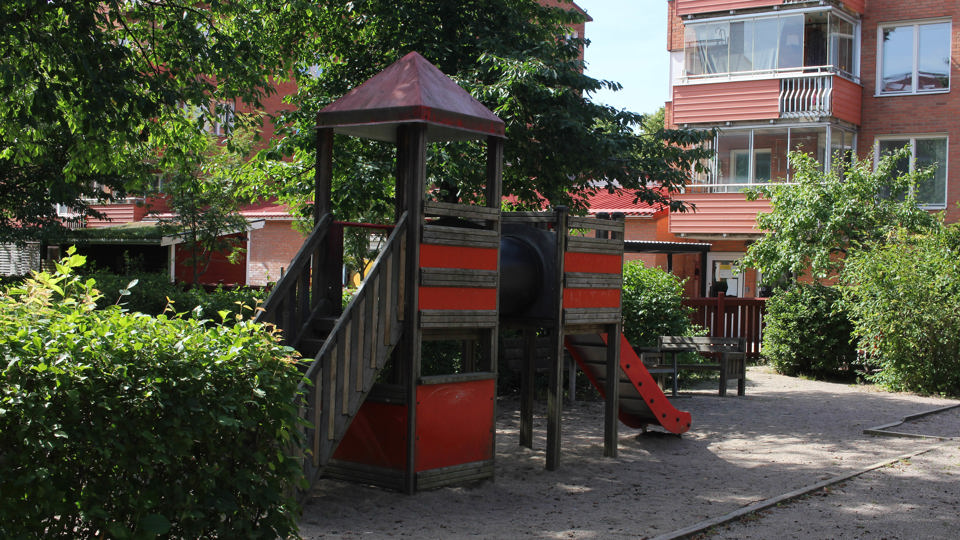 En stor röd klätterställning i ett bostadsområdes grönskande innergård framför förskolan Ballongen som ligger i ett flerfamiljshus i tegel.