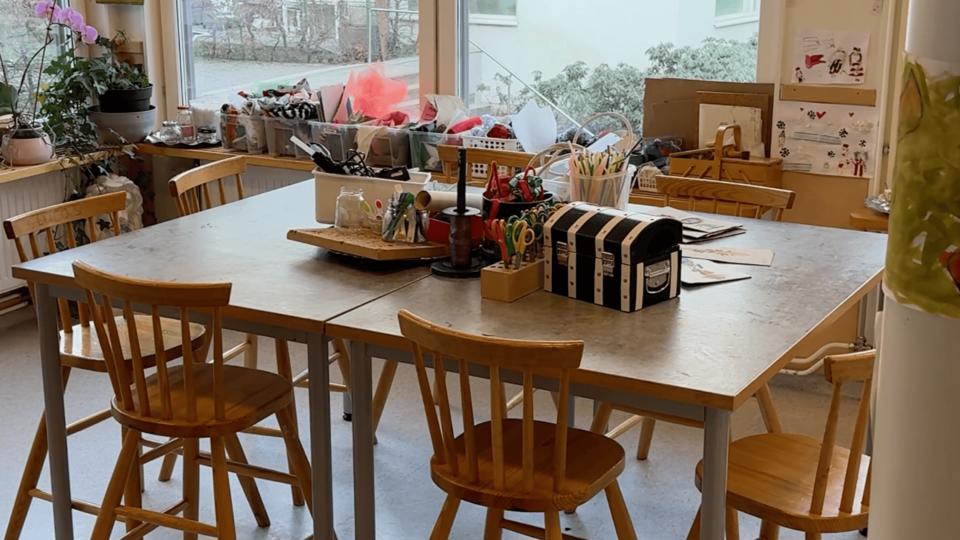 Inomhus i ett förskoleklassrum med ett stort bord, stolar och pysselmaterial på bordet samt hyllor.