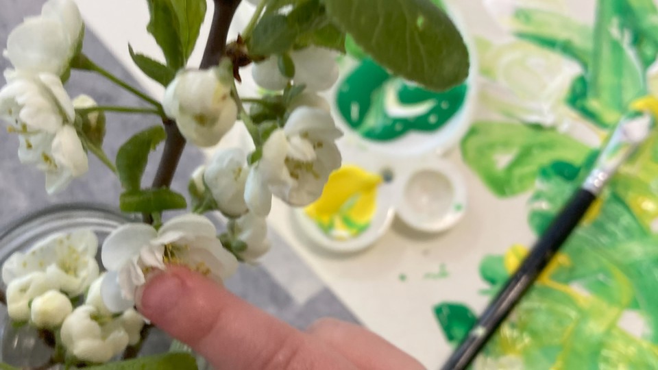En barnhand som håller i en blomsterstängel. På bordet i bakgrunden ligger ett papper, pensel och vattenfärg. Foto. 