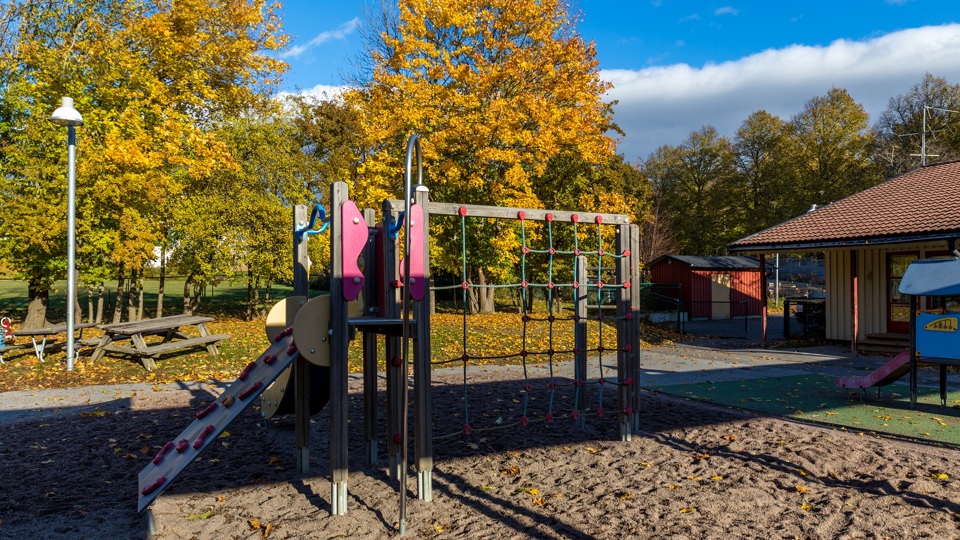 Byggnad med gård framför med fallna höstlöv, träd, sittplatser, sandlåda, klätterställning och rutschkana.