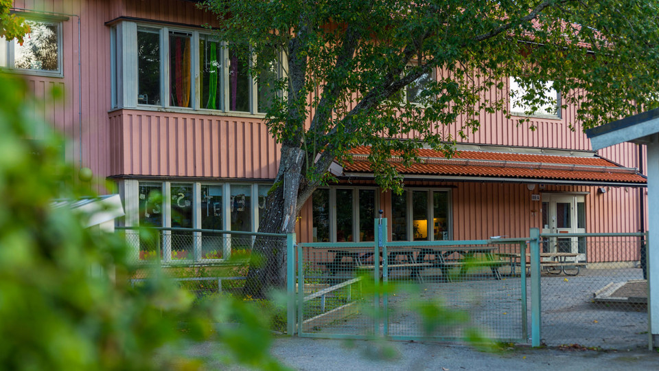 Staket och grind in till förskolebyggnad. Ett stort träd inramar entrén.
