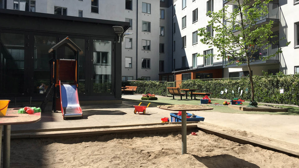 Utemiljö med gård, sandlåda och rutschkana. Ljusa flerbostadshus i bakgrunden.
