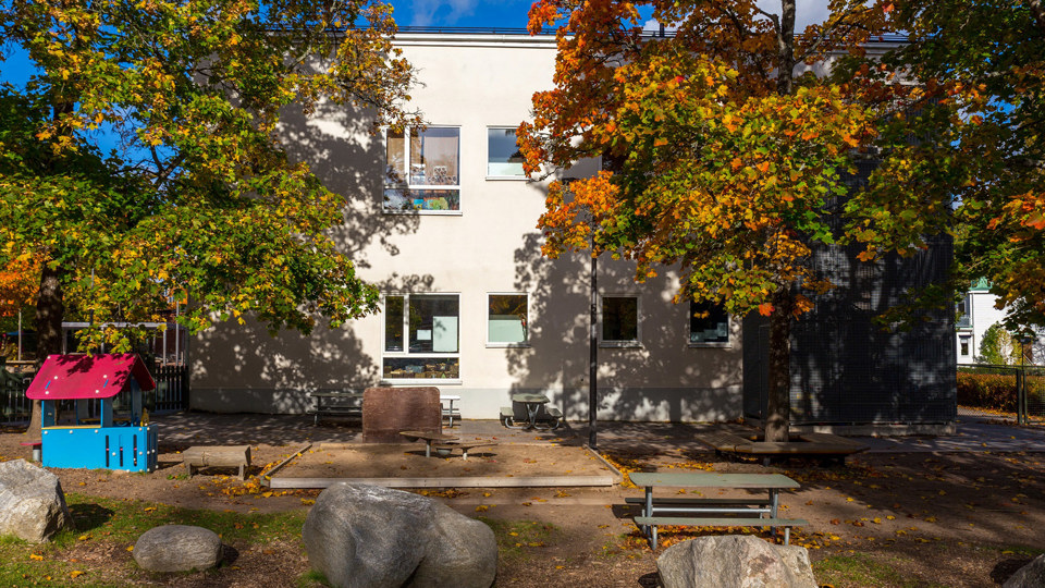 Lekyta med sandlåda och lekredskap framför ett hus i två plan. Grönskande träd.