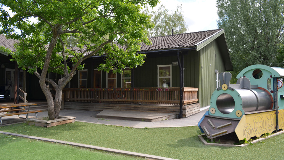 Bild på förskolans gröna träfasad, sandlåda och lekutrustning. På vår innegård finns det gott om plats, material och lekutrustning för aktiva lekar.