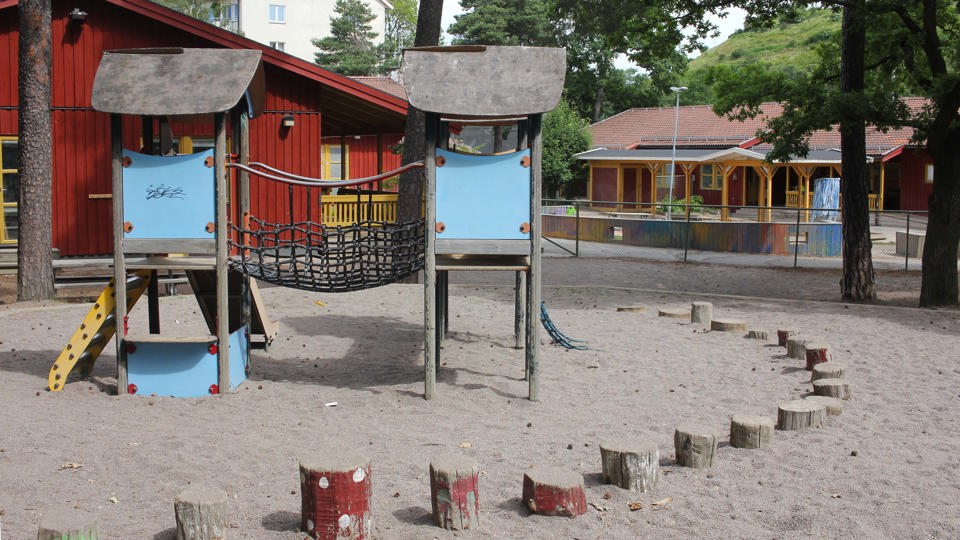 En klätterställning i trä, blått och rött står på en yta täckt med sand och runt området finns målade stubbar. I bakgrunden skymtas två röda enplanshus.