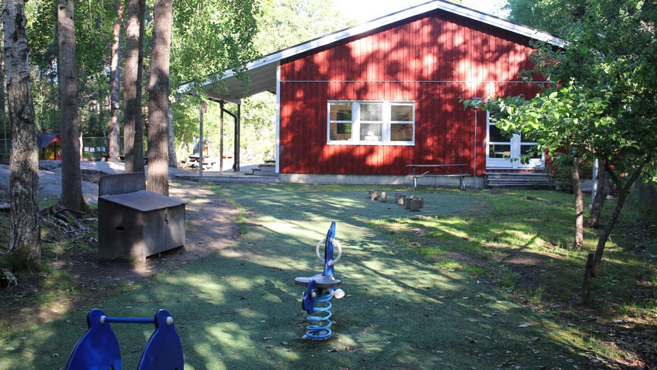 Solen lyser på ett rött enplanshus i ett grönt skogsparti. Framför huset finns två blå gunghästar.