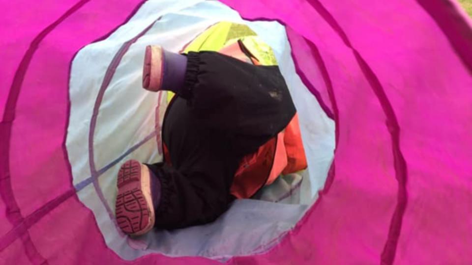 Ett barns ben syns i en färgglad lek-tunnel, med svarta byxor och lila skor.