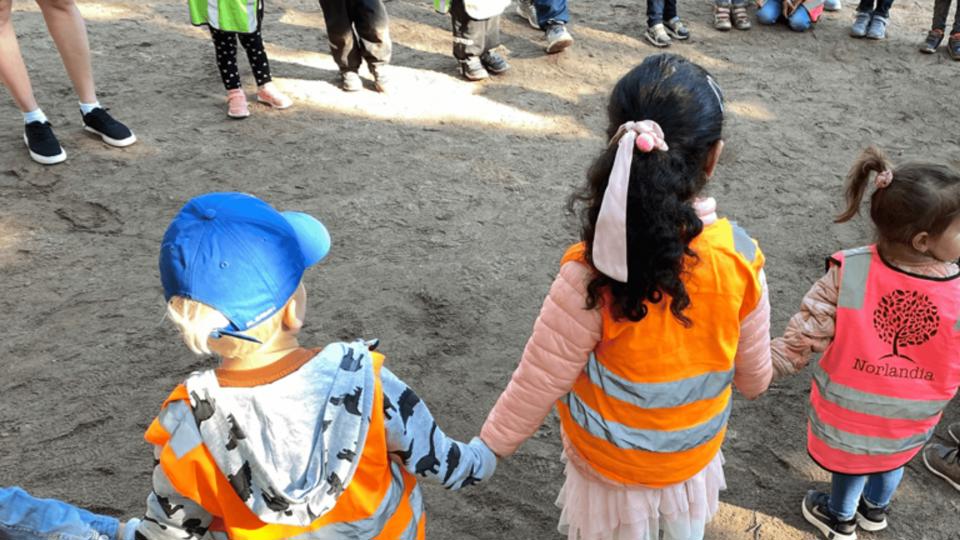 Förskolebarn håller varandra i händerna utomhus i en cirkel och har på sig reflexvästar.