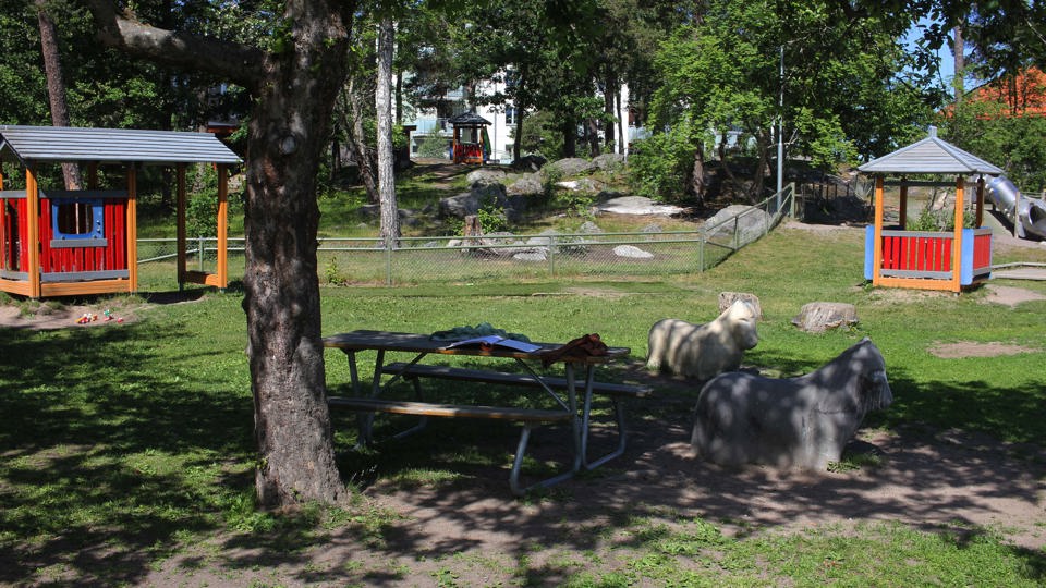 Två röda lekhus, en sittyta och konstgjorda getter på en grönskande förskolegård.