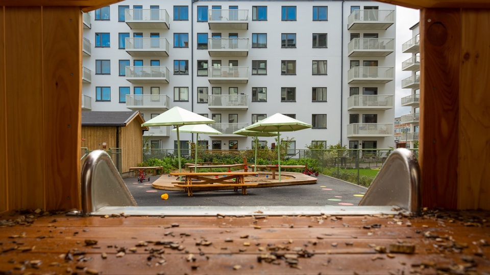 Utsikt över flerbostadshusets innergård från klätterhuset. Man ser sandlåda, lekobjekt, buskar och solskydd.