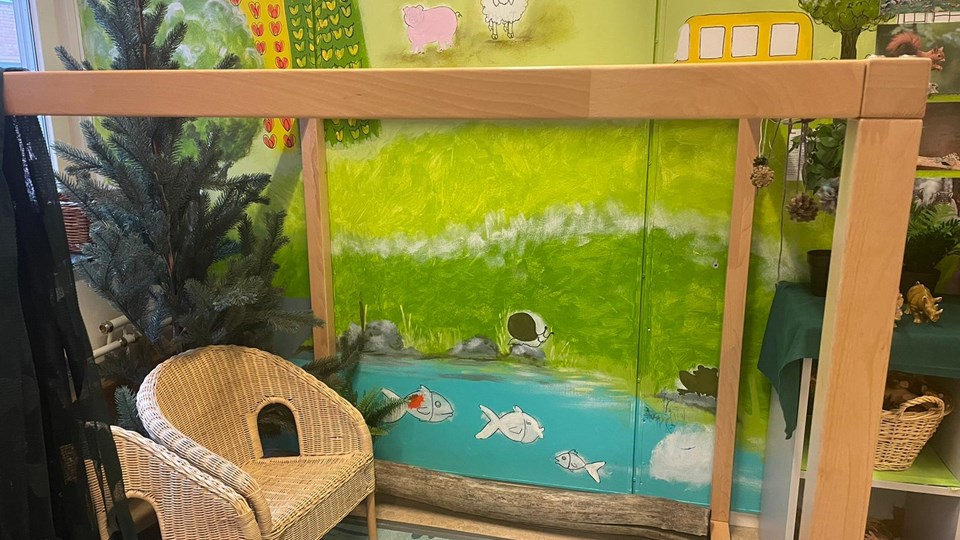 Två små stolar och hyllor med plastdjur som står framför en väggmålning med natur, djur och fiskar i en fiskdamm.