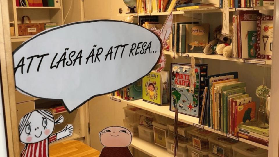 Pappfigurer som uppmuntrar till läsning står framför bokhyllor, foto.