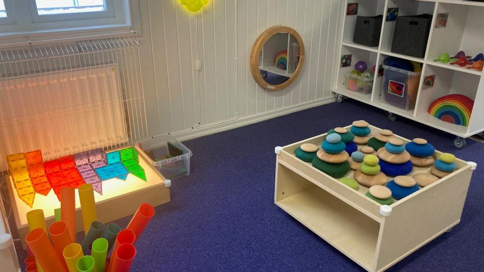 Ett rum för lek med ljusbord, färgade byggklossar, stapelleksaker och en spegel på väggen.