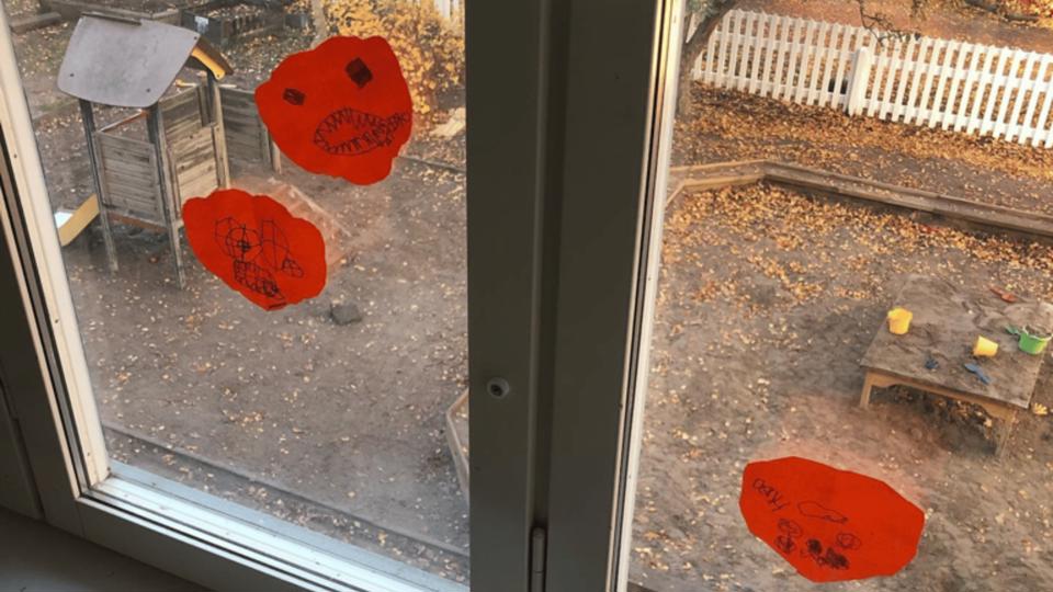 Teckningar på ett fönster i förskolans inomhusmiljö med utsikt mot sandlåda och lekplats.