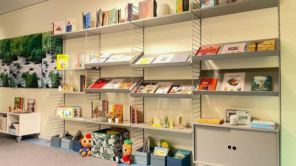 Barnböcker visas på grå vägghyllor i ett bibliotek med lådor, spel och mjukisdjur på golvet.