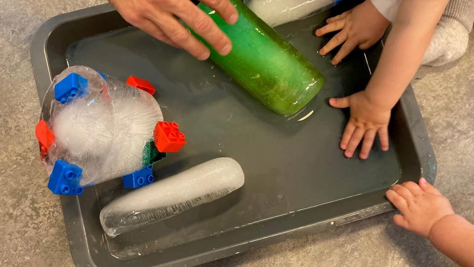 Barn och en vuxen leker med is i en vattenbalja på golvet, med frusna flaskor och leksaker.