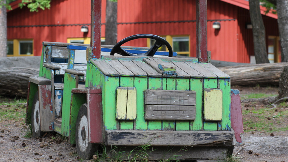 En grön stor leksaksbil i trä, för små barn, står på en skogslik förskolegård framför ett rött enplanshus.