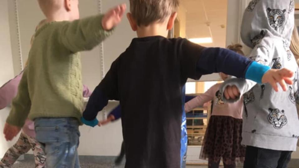 En grupp förskolebarn leker i en cirkel inomhus och håller varandras händer.