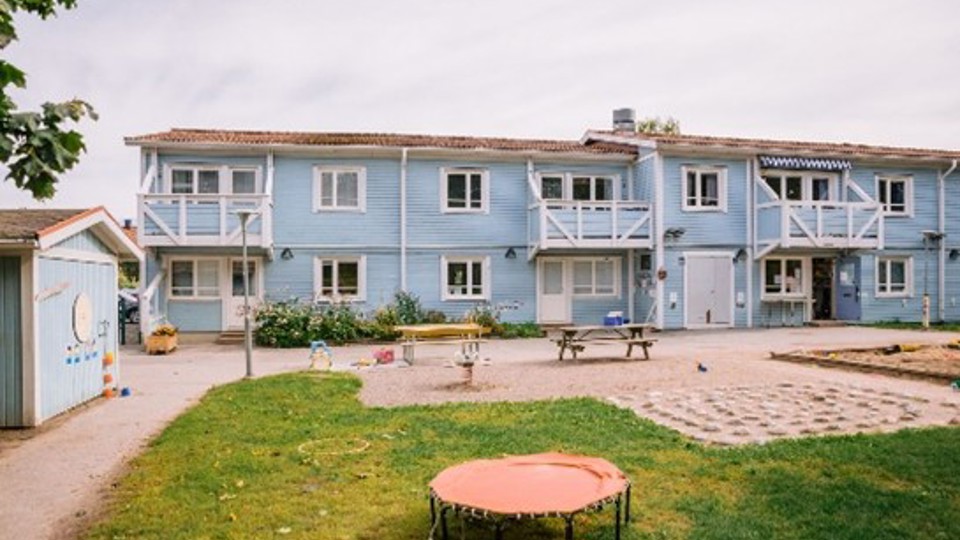 Ljusblått trähus i två våningar och en skolgård med sandlåda och sittplatser.