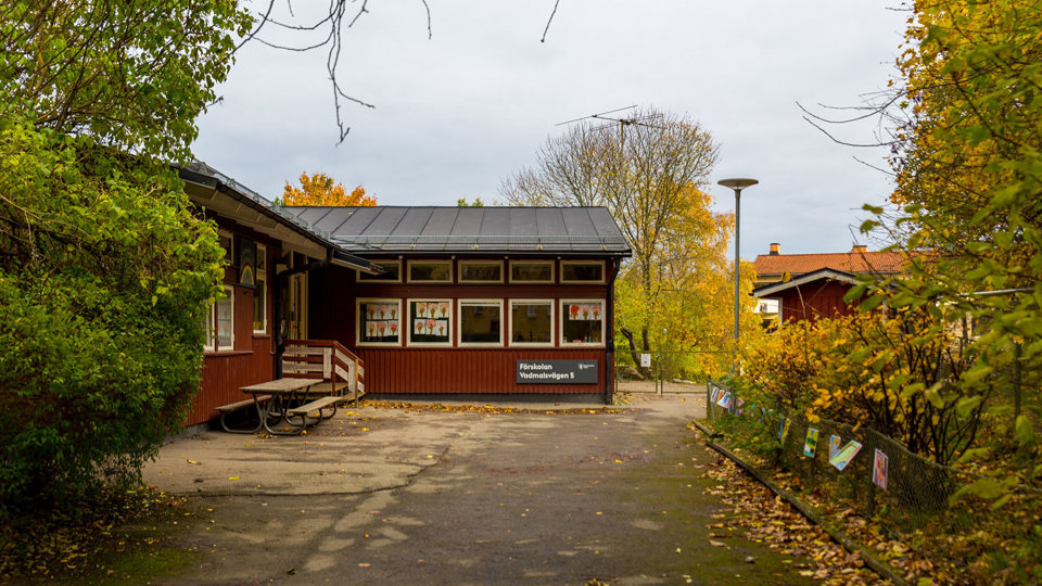 Röd träbyggnad omgiven av grönska och gulnande löv, foto.