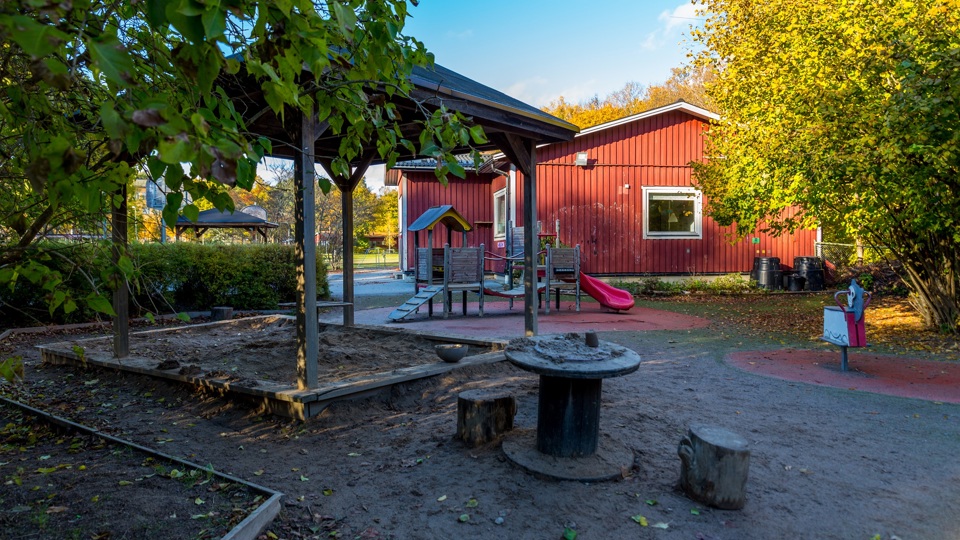 Byggnad med sandlåda, träd, klätterställning och andra lekobjekt på gården framför sig.
