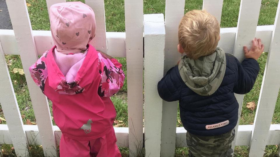 Barn i regnkläder tittar genom ett vitt staket.