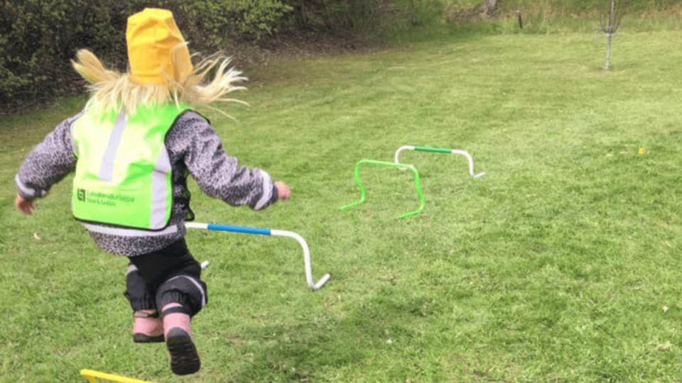 Ett barn hoppar över hinder på en gräsmatta under en utomhusaktivitet, med reflexväst och gul mössa.