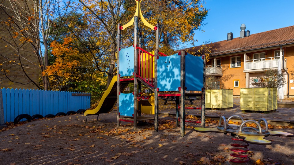 Gård med klätterställning, sandlåda och träd framför en byggnad