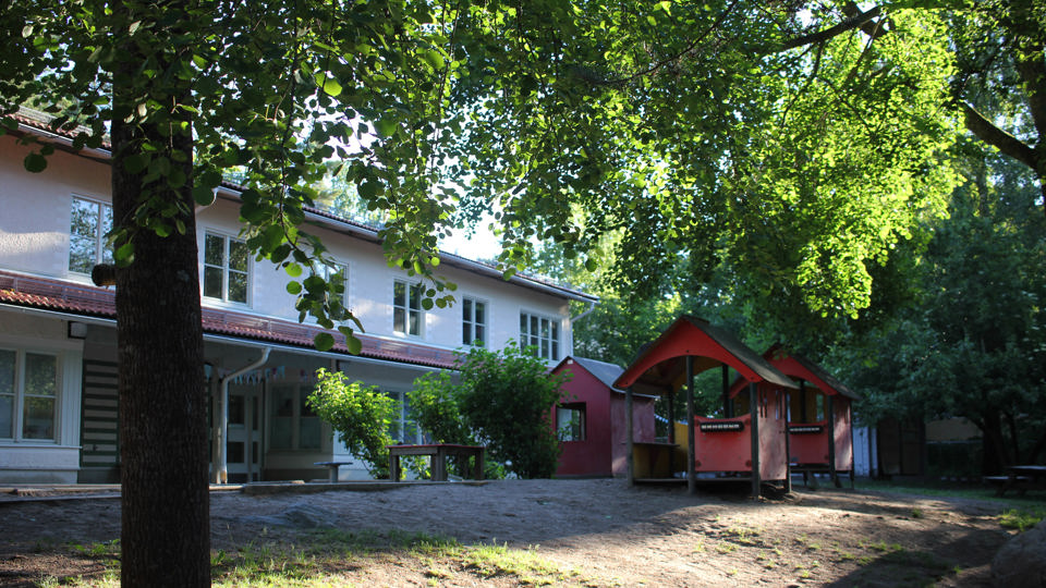 Förskolan Fyrbåken ligger på en grön kulle på en grönskande gård. Förskolan är ett tvåplanshus i en putsad naturvit yta med röda tegelpannor.
