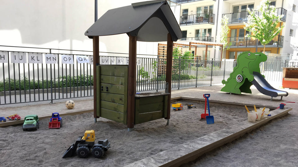 Utemiljö med lekytor, sandlåda och rutschkana. Ljusa flerbostadshus i bakgrunden.