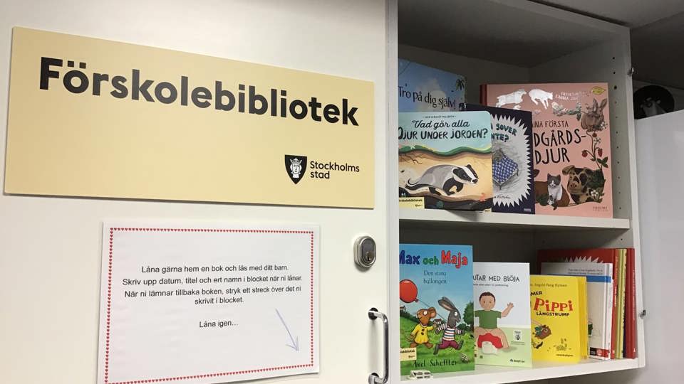 Skylt med texten ”Förskolebibliotek” på ett skåp med barnböcker och information om att låna hem böcker.