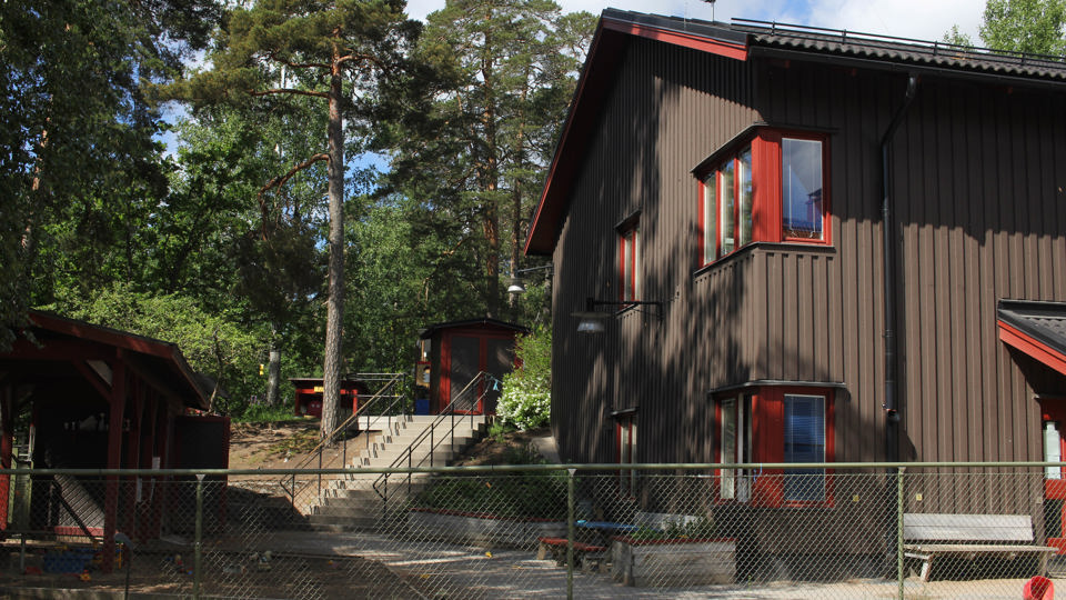 Ett tvåplanshus i brunt trä, röda fönsterkarmar och röd dörr står bakom ett grönt staket. Intill huset syns ett förråd och en trappa leder till en annan del av gården där det finns ett rött lekhus.