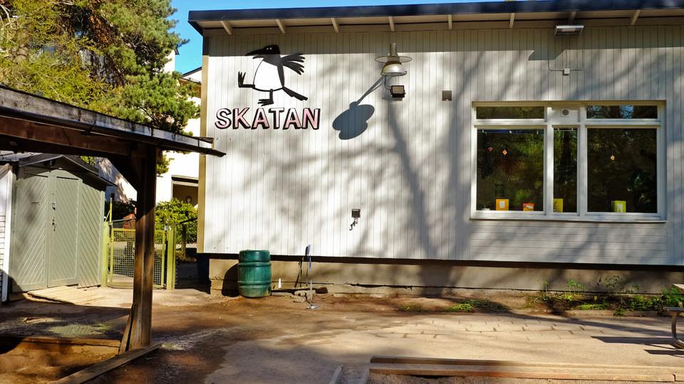 Vit byggnad med skylten ”Skatan” och fågellogga i ett soligt skogsområde, med sandlåda och gård i förgrunden.
