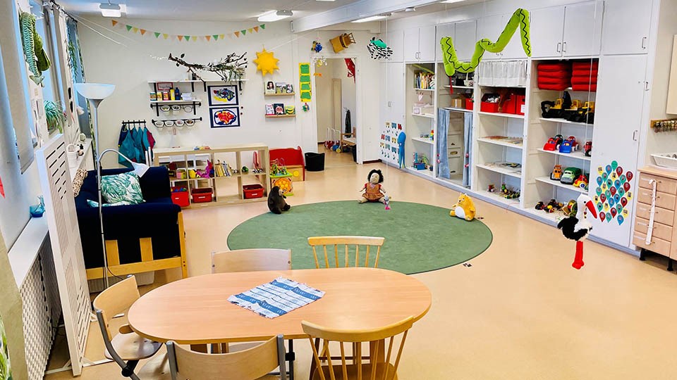 Rum med bord, stolar, grön matta, soffa och lekmaterial för små barn.