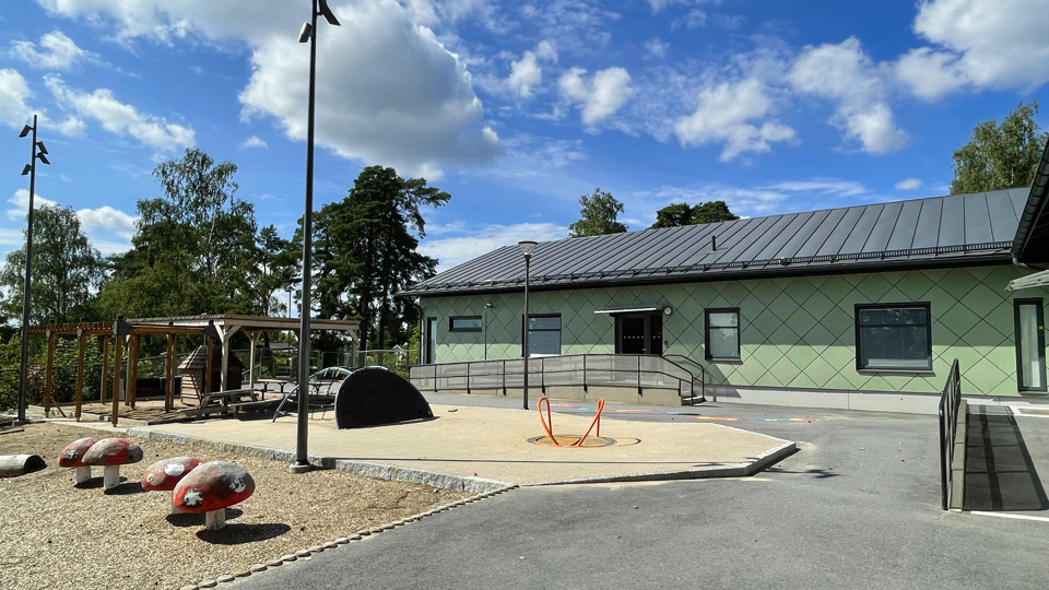 Hus med lekplats, sandlåda och asfalt på gården. Träd syns i bakgrunden.