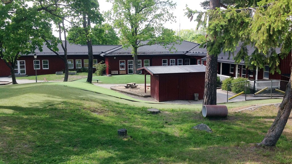 Lummig gård med gräsytor för lek, med ett rött enplanshus i bakgrunden.