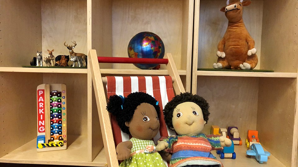 Foto på två dockor som sitter i en solstol framför en hylla med leksaker.