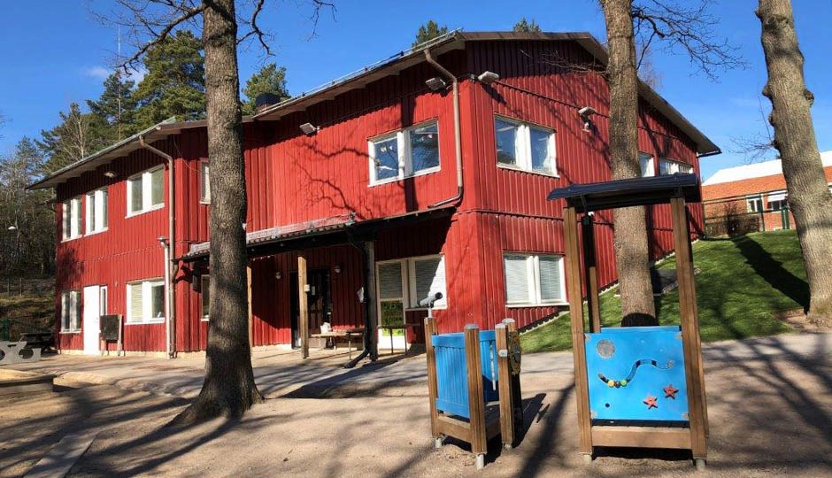 Byggnad i två plan med röd fasad i trä. Stora trädstammar, sandlåda och blå lekutrustning i förgrunden.