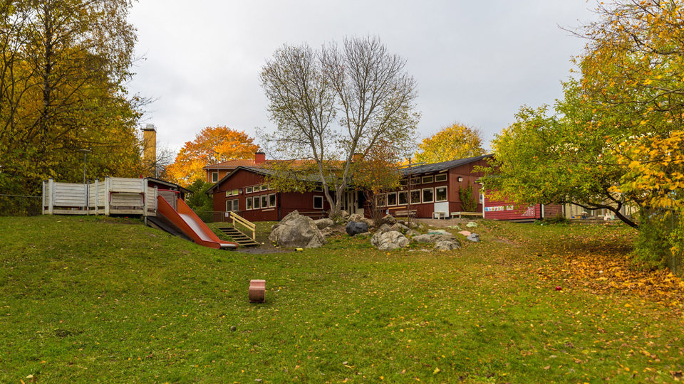 Rött trähus omgivet av grönska och solbelysta träd med gula blad, foto.