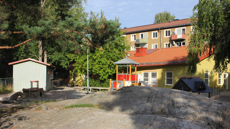 Förskolan Ankaret, ett gult enplanshus med en gård. På gården syns en gul och röd klätterställning och småhus, berg och träd. Bakom gården och förskolan synd ett flerfamiljshus i brunputsad fasad.