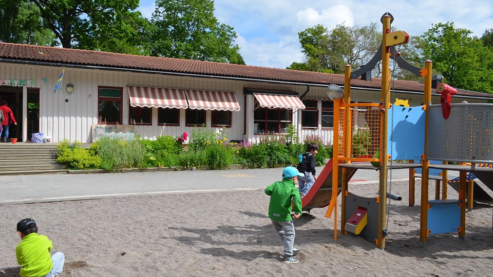 Bild på förskolans vita träfasad och lekande barn. Den stora innergården erbjuder fina lekytor, lekutrustning och material för aktiva lekar.