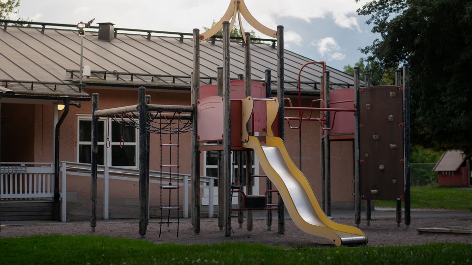 Förskolans lekplats med klätterställning och gul rutschkana framför byggnaden, omgiven av gräs och träd.