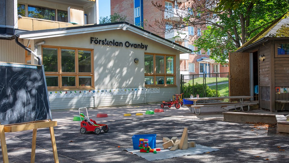 Förskolegård med sandlåda och pysselbord, samt asfalt med leksaksbilar, trehjulingar, klossar och staffli.