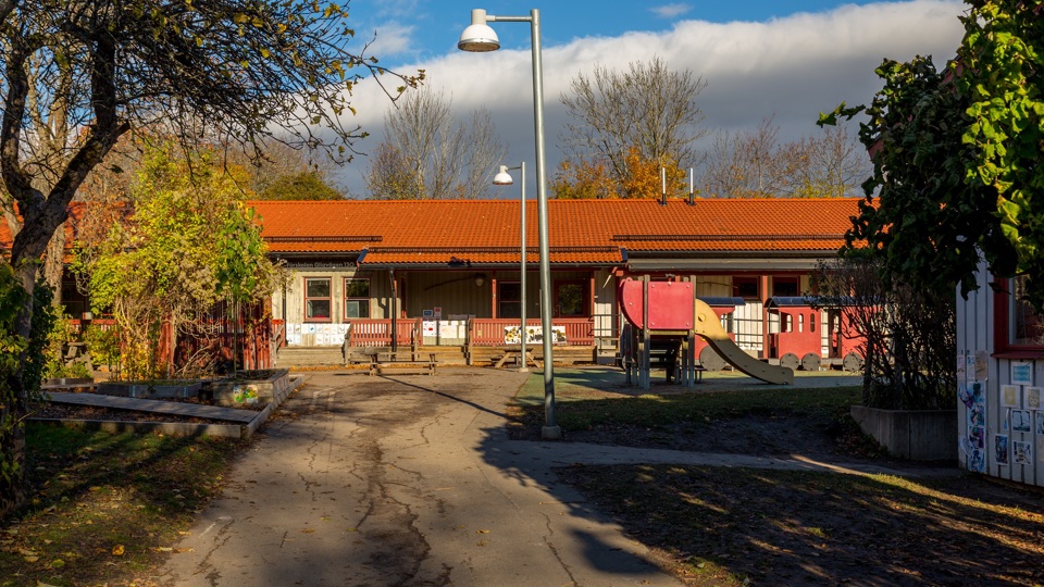 Byggnad med asfalt, buskar träd, klätterställning och lektåg på gården framför sig.