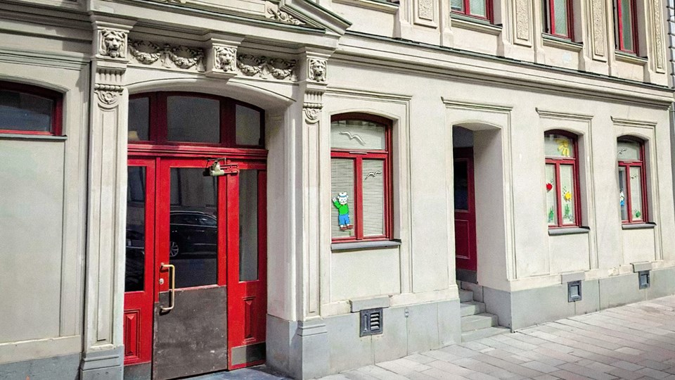 Entré från gatan till förskolan Flaggskepet. Stenbyggnad med röd dörr och fönster som är smyckade med olika pynt. Foto.